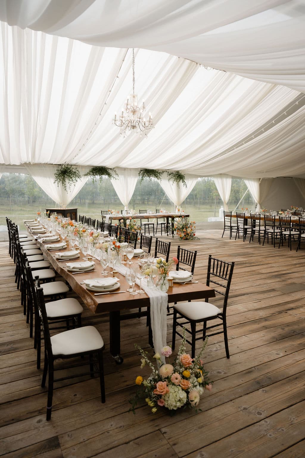 Long harvest table set with peach and white florals under a white draped tent, chandelier and greenery overhead