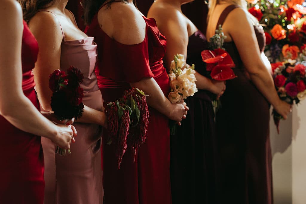Bridesmaids in red, blush and burgundy dresses, each holding a single-variety bouquet of dahlias, amaranthus, lisianthus or anthurium
