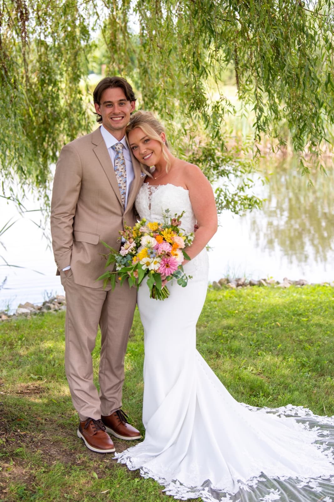 Newlyweds under a willow at the water's edge, the bride holding a bright hand-tied bouquet