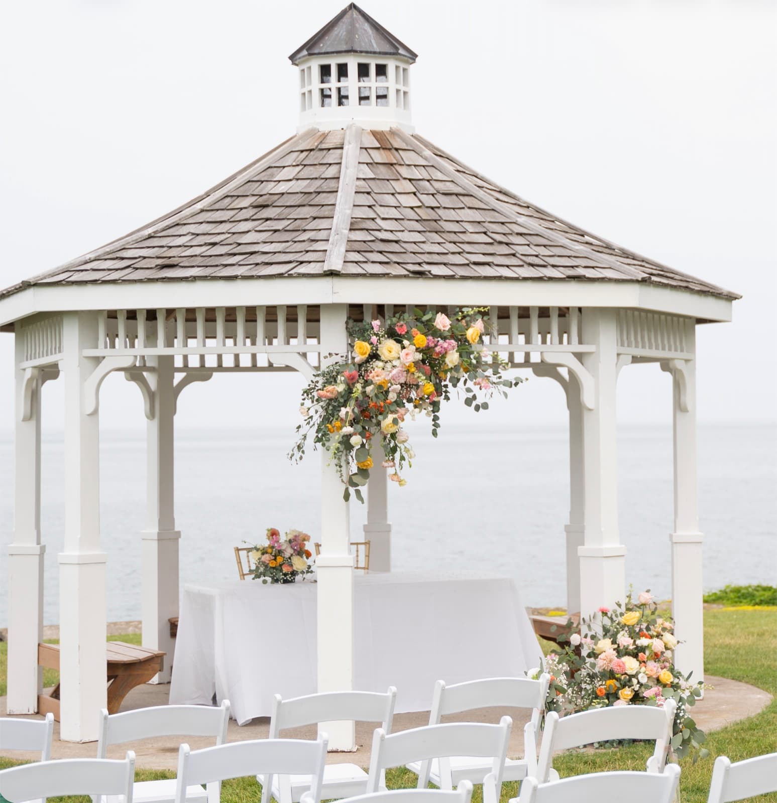 A white lakeside gazebo with a floral spray on its post, a welcome table set beneath and an arrangement beside the chairs