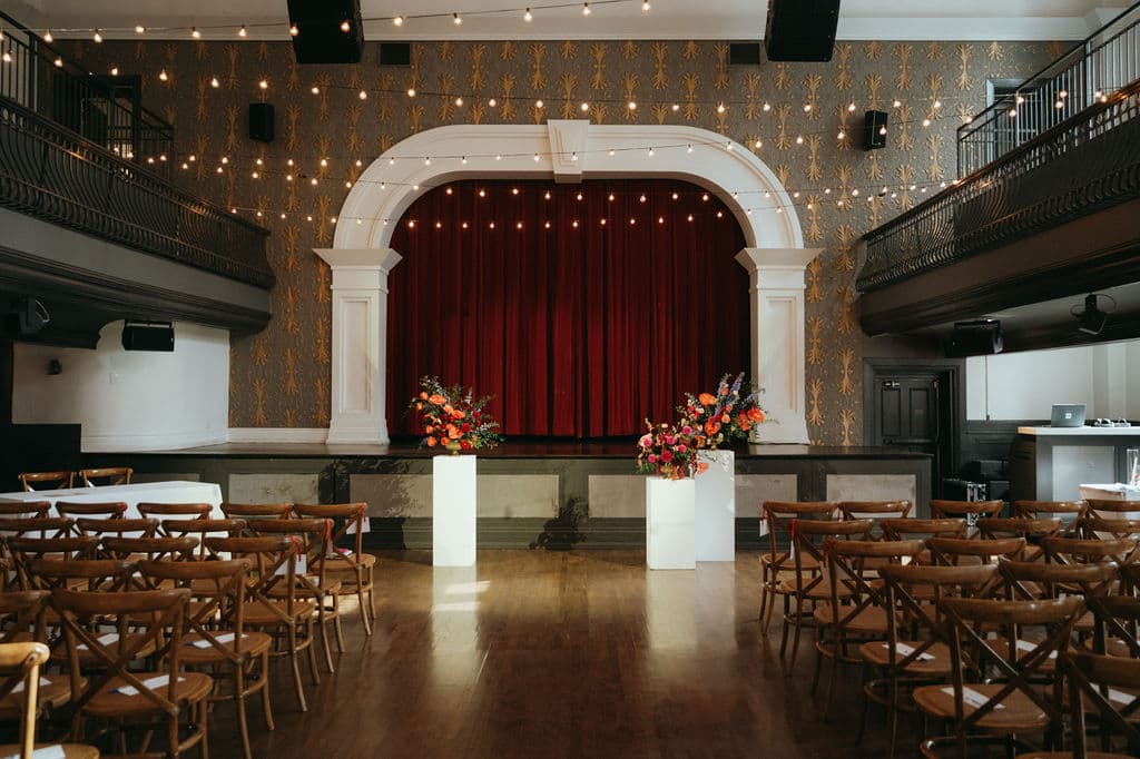 Two pedestal arrangements of orange and red blooms flanking a theatre stage with a red velvet curtain