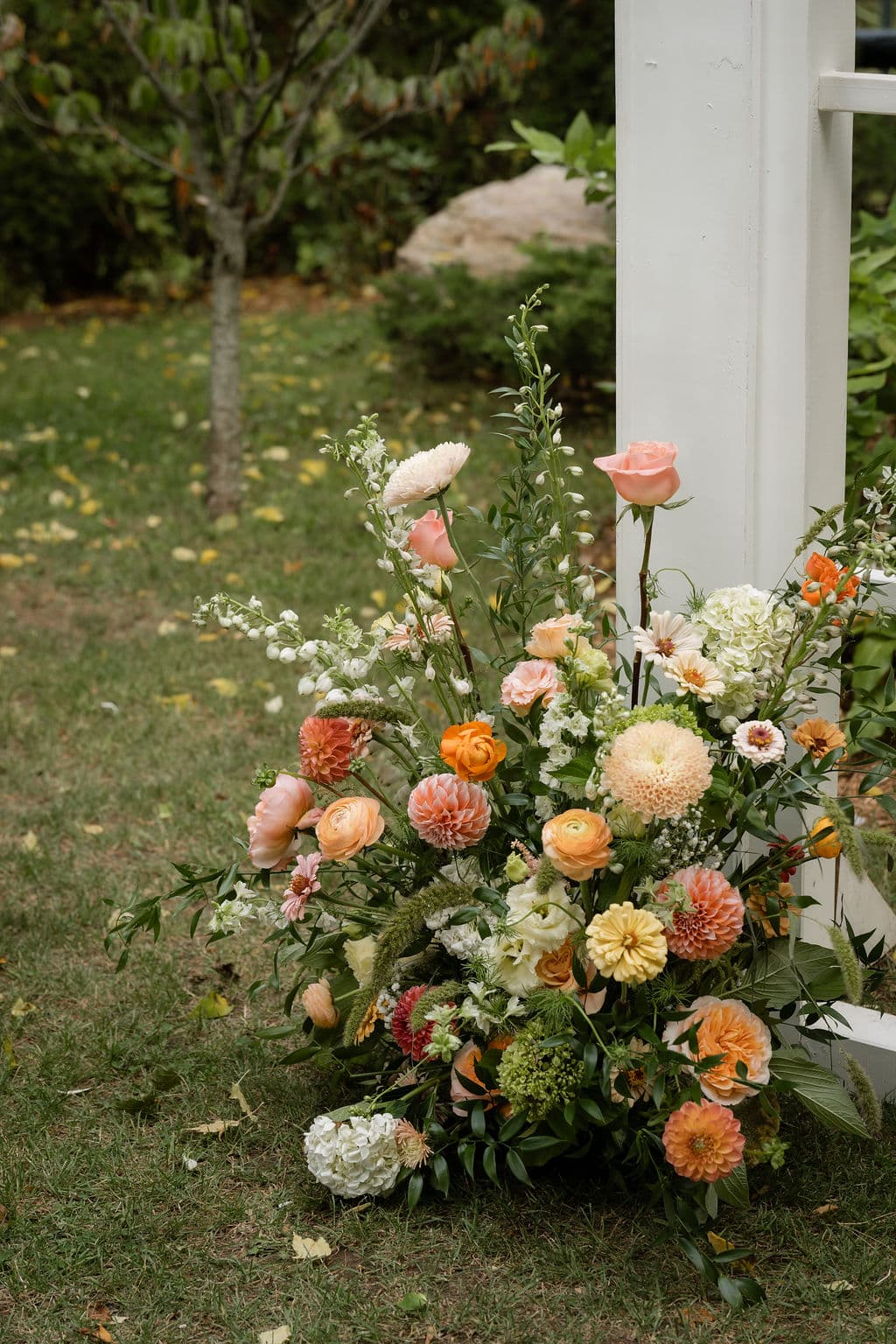A garden-style arrangement of peach dahlias, roses and zinnias at the base of a white arch post