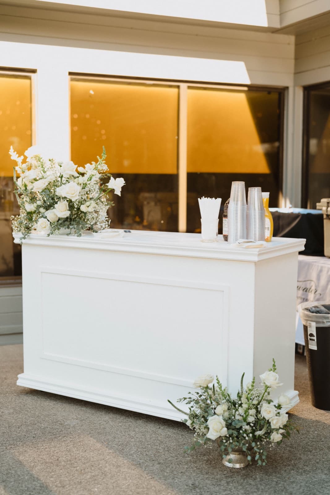 A white bar with a spray of white roses and greenery on its corner and an arrangement at its base