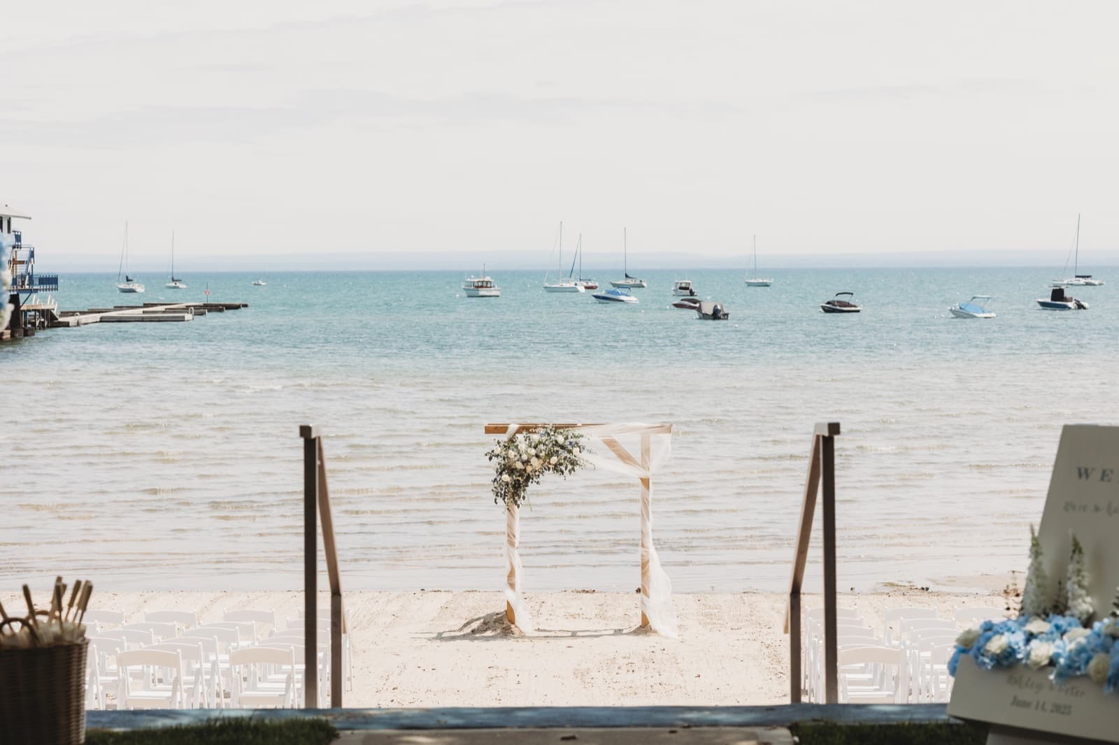 A ceremony arch topped with greenery on a beach, sailboats moored on the water beyond