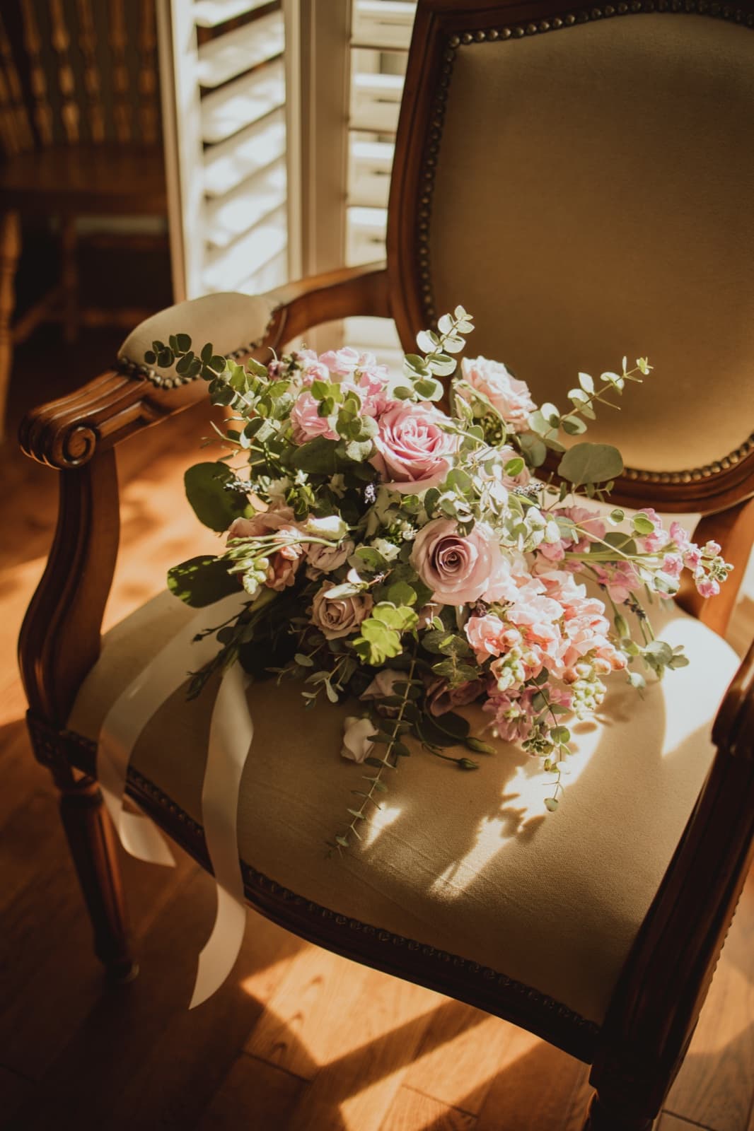 A pastel bridal bouquet of roses and eucalyptus resting on an antique armchair in window light
