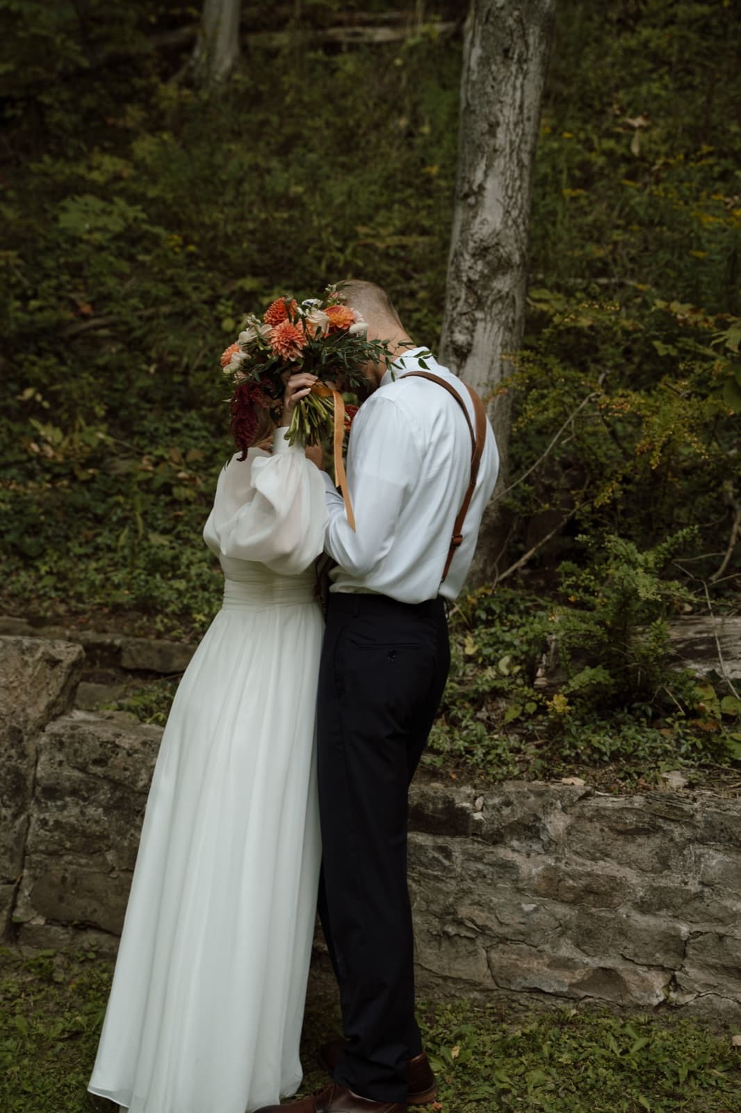 Newlyweds stealing a kiss behind a bouquet of orange dahlias at the edge of the woods