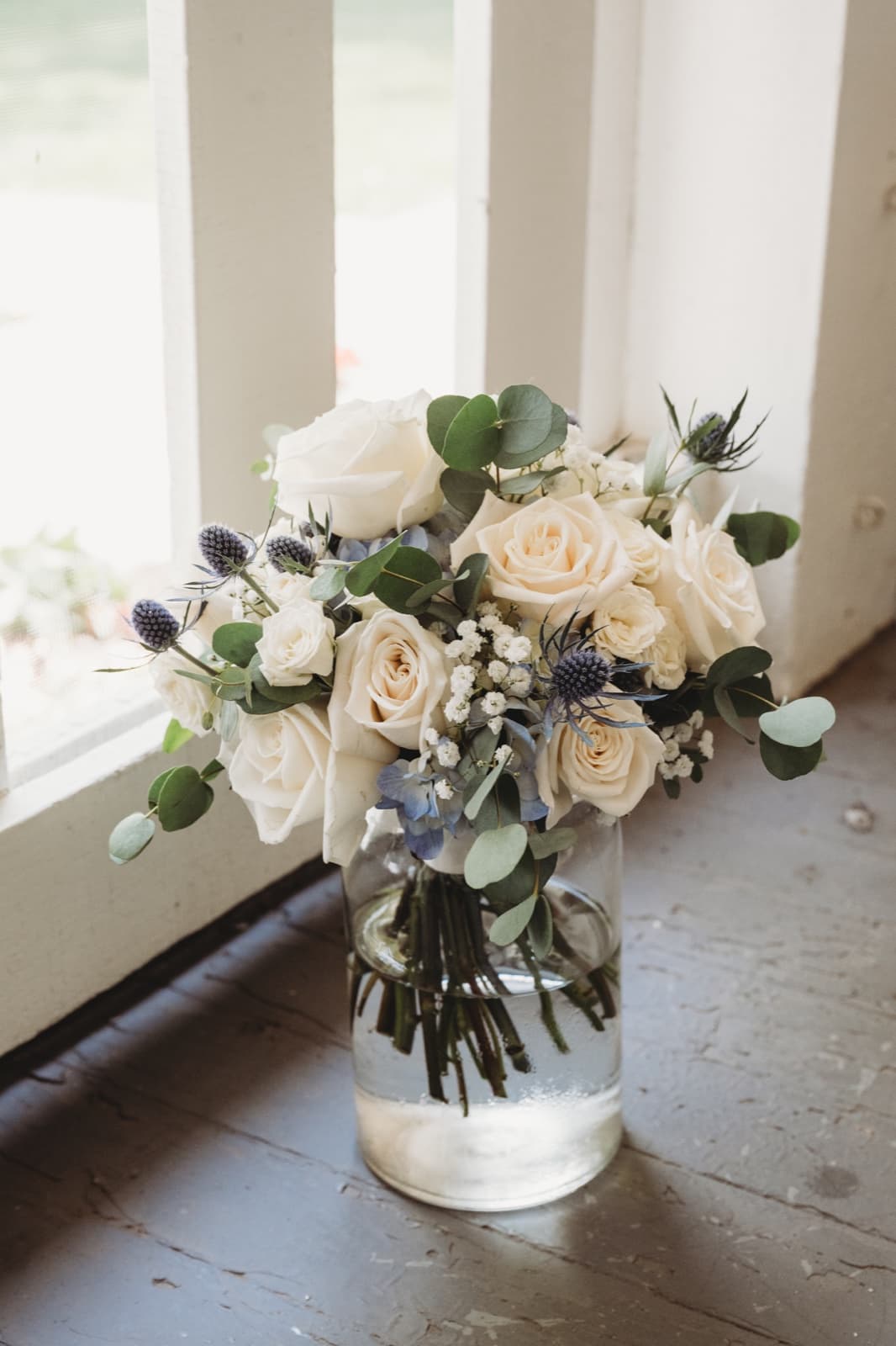A bouquet of white roses, blue sea holly and eucalyptus in a glass jar beside a window