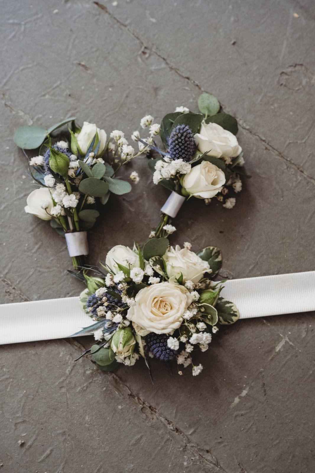 Two boutonnieres and a wrist corsage of white spray roses, sea holly and baby's breath laid out on a stone floor