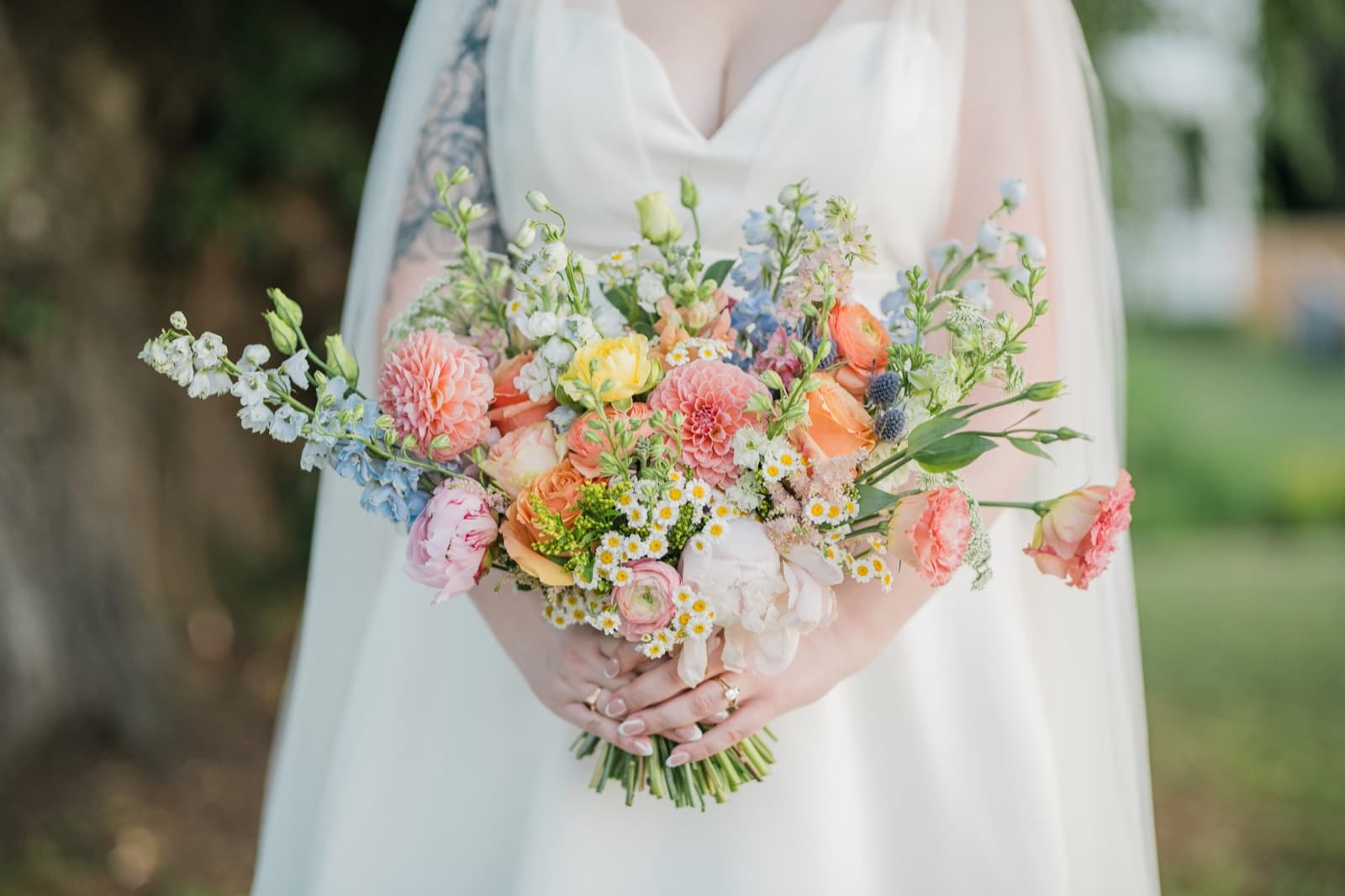 Bride holding a colorful garden-style bouquet of coral dahlias, peach roses, ranunculus, chamomile and delphinium against a white dress