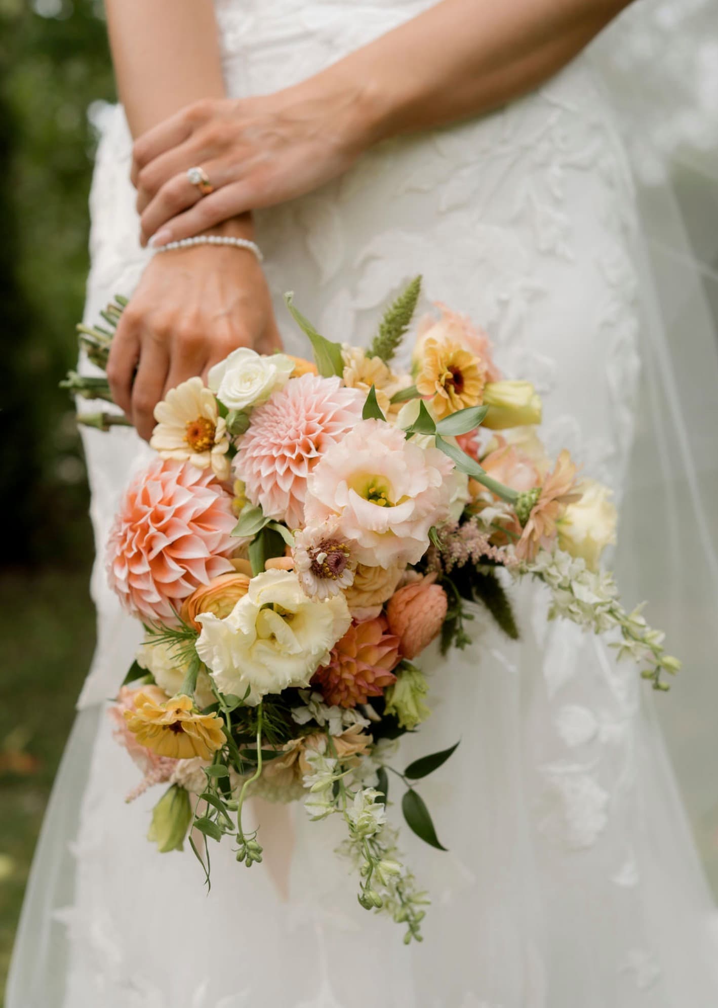 Close-up of a bride holding a hand-tied bouquet of peach dahlias, lisianthus, zinnias and ranunculus against a white lace dress