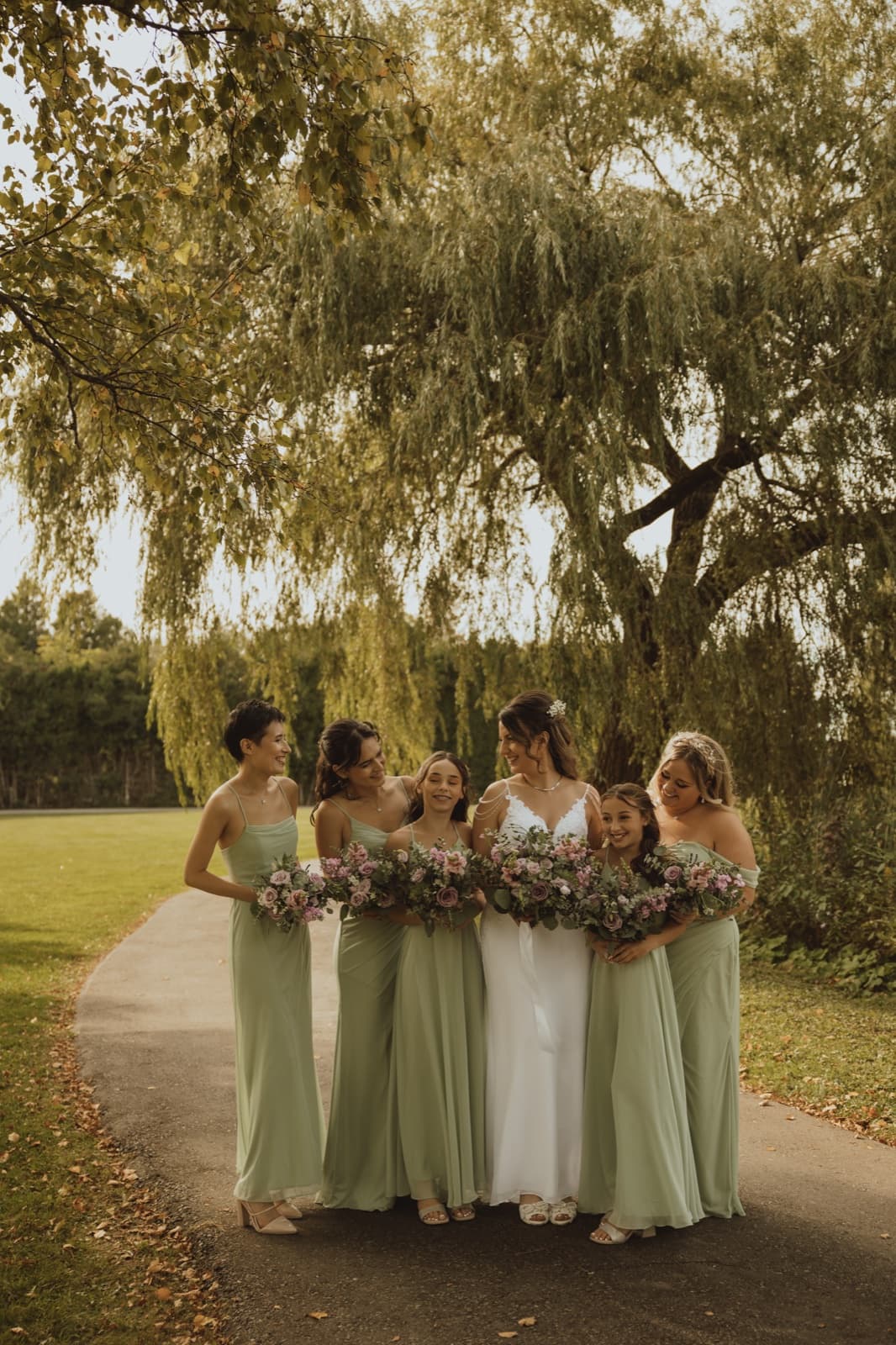A bride and her bridesmaids in sage dresses holding dusty pink bouquets under a willow tree