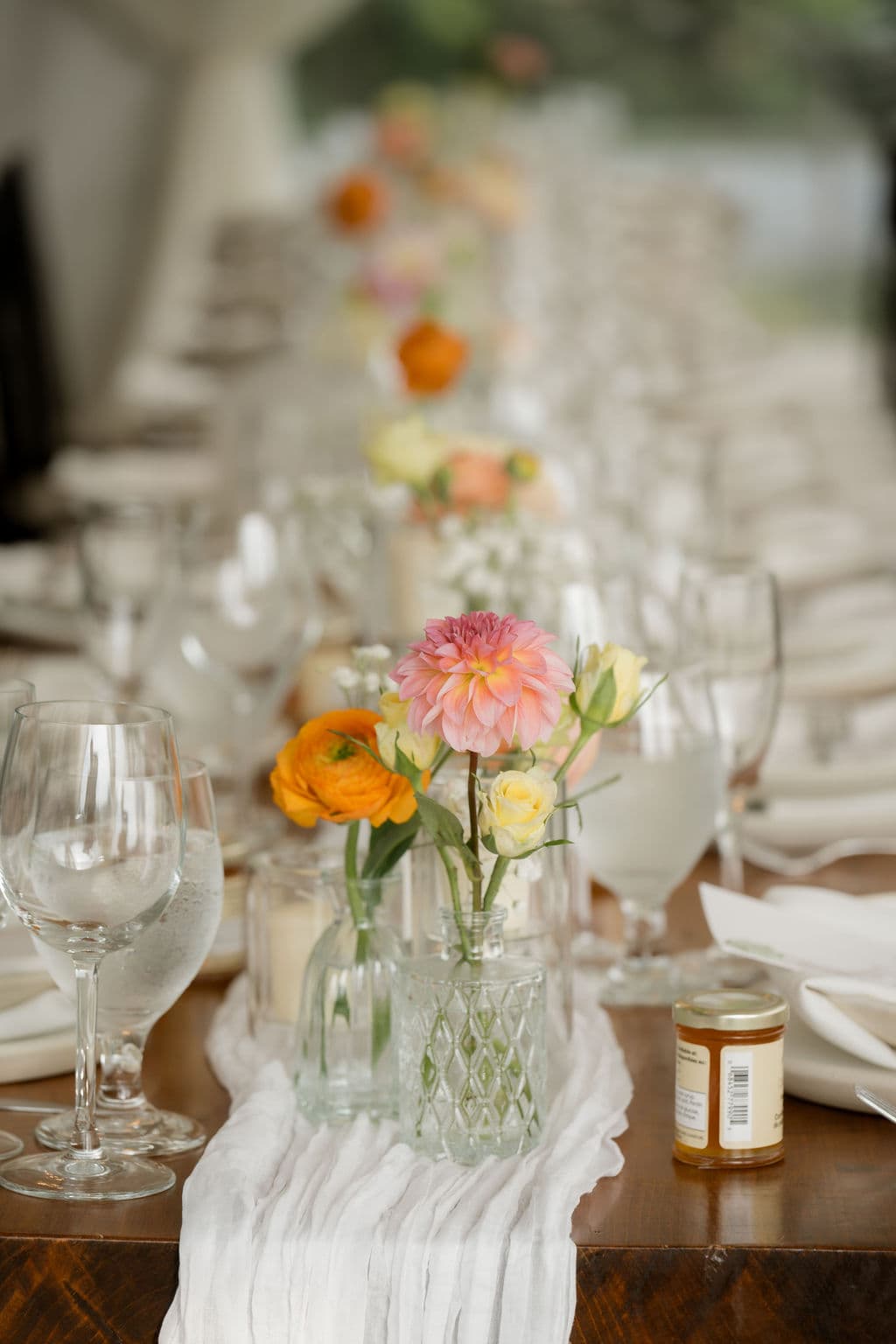 Bud vases of dahlias, ranunculus and roses running down a harvest table with a gauze runner