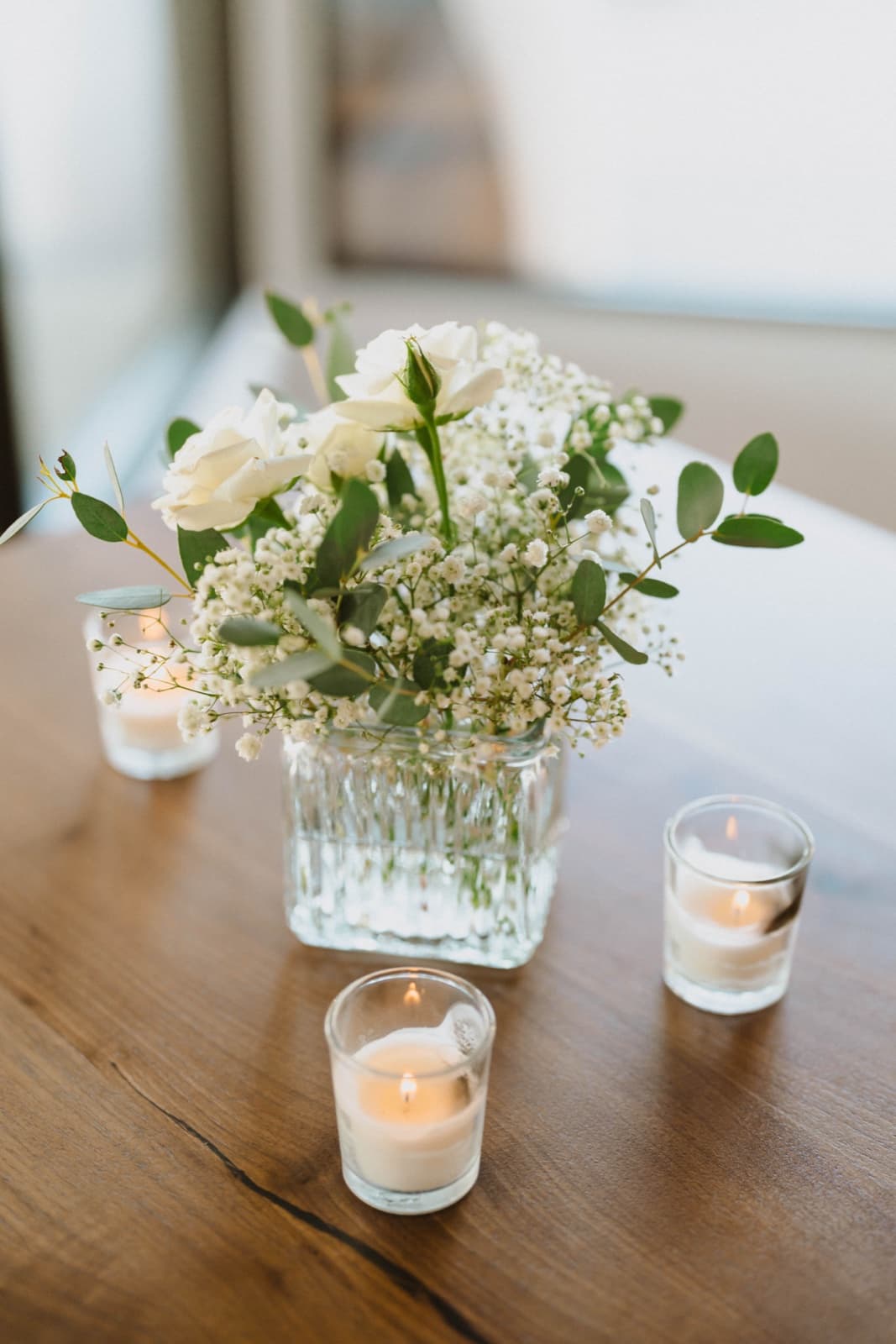 A glass bud vase of white roses, baby's breath and eucalyptus ringed by votive candles on a wood table