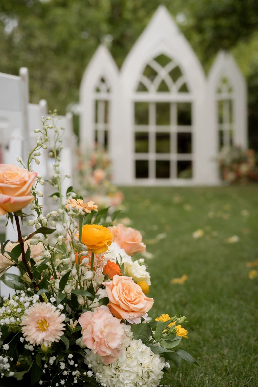 Peach and orange aisle arrangement of roses, ranunculus and hydrangea beside white ceremony chairs, with a white chapel-arch backdrop on the lawn