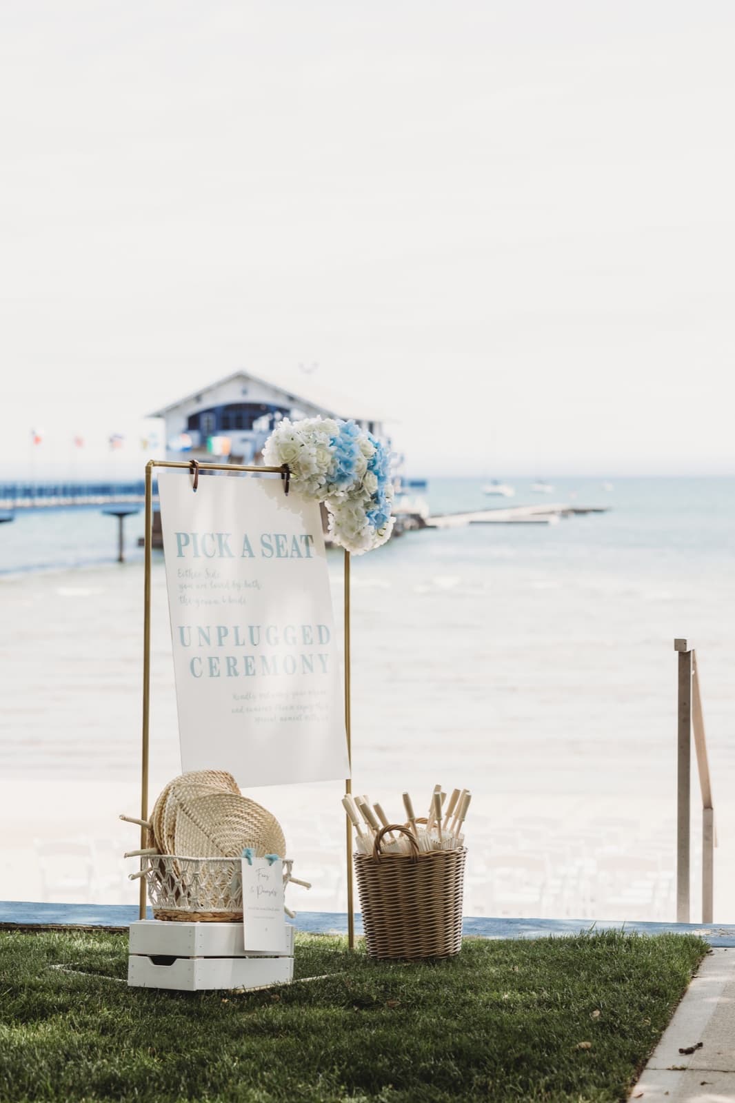 A lakeside ceremony sign topped with blue and white blooms, fans and parasols waiting in baskets