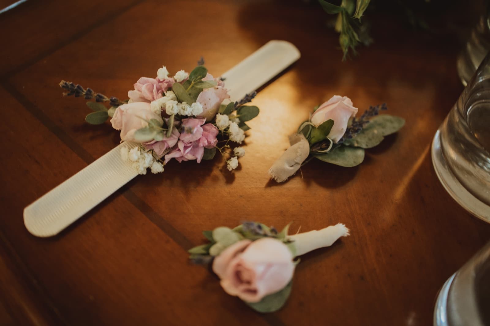 A wrist corsage and two blush rose boutonnieres with lavender and baby's breath laid out on a wooden table