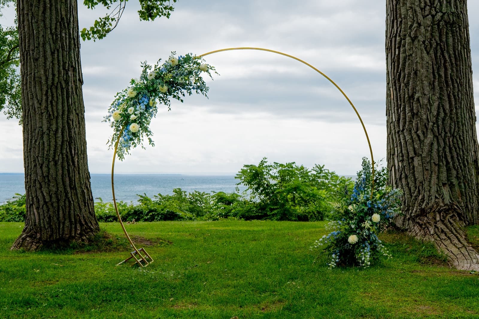 Gold circular wedding arch dressed with blue and white florals and greenery, standing between two trees on a lakeside lawn