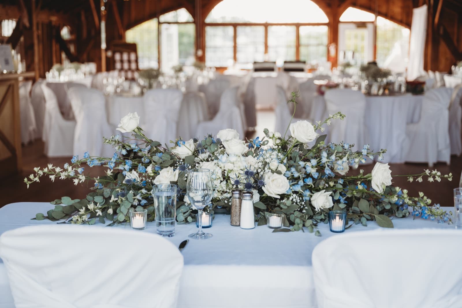 A long low arrangement of white roses, blue delphinium and eucalyptus with candles on a head table