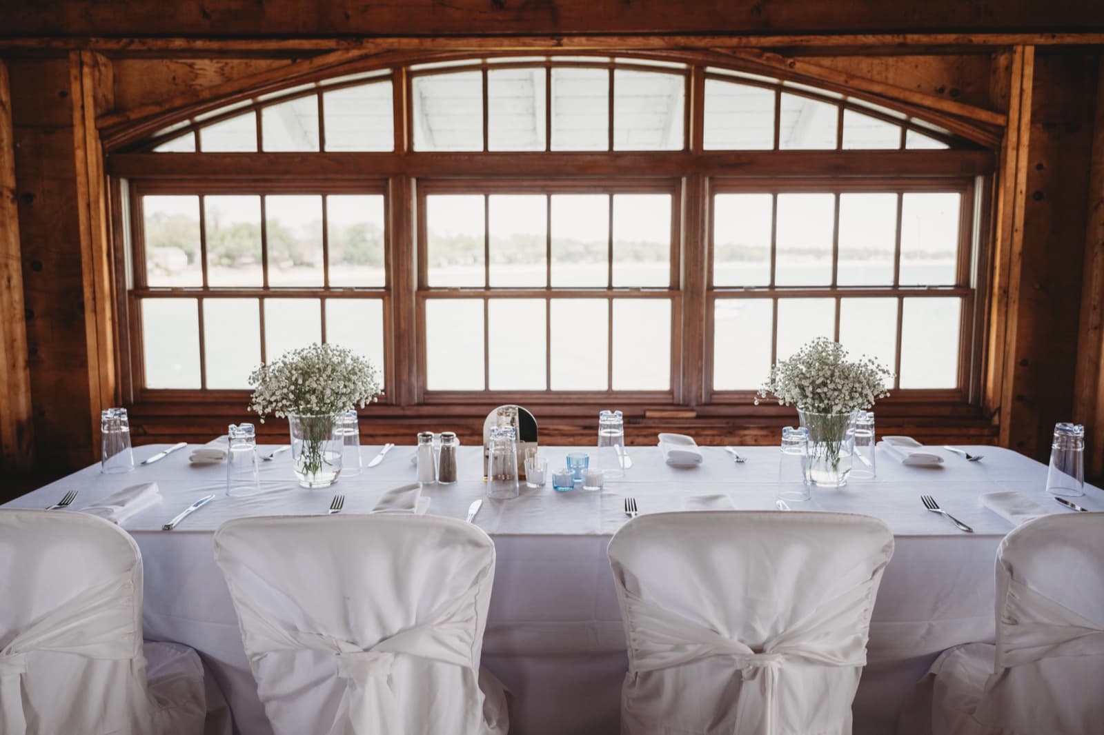 A white head table set before lakefront windows with two vases of baby's breath