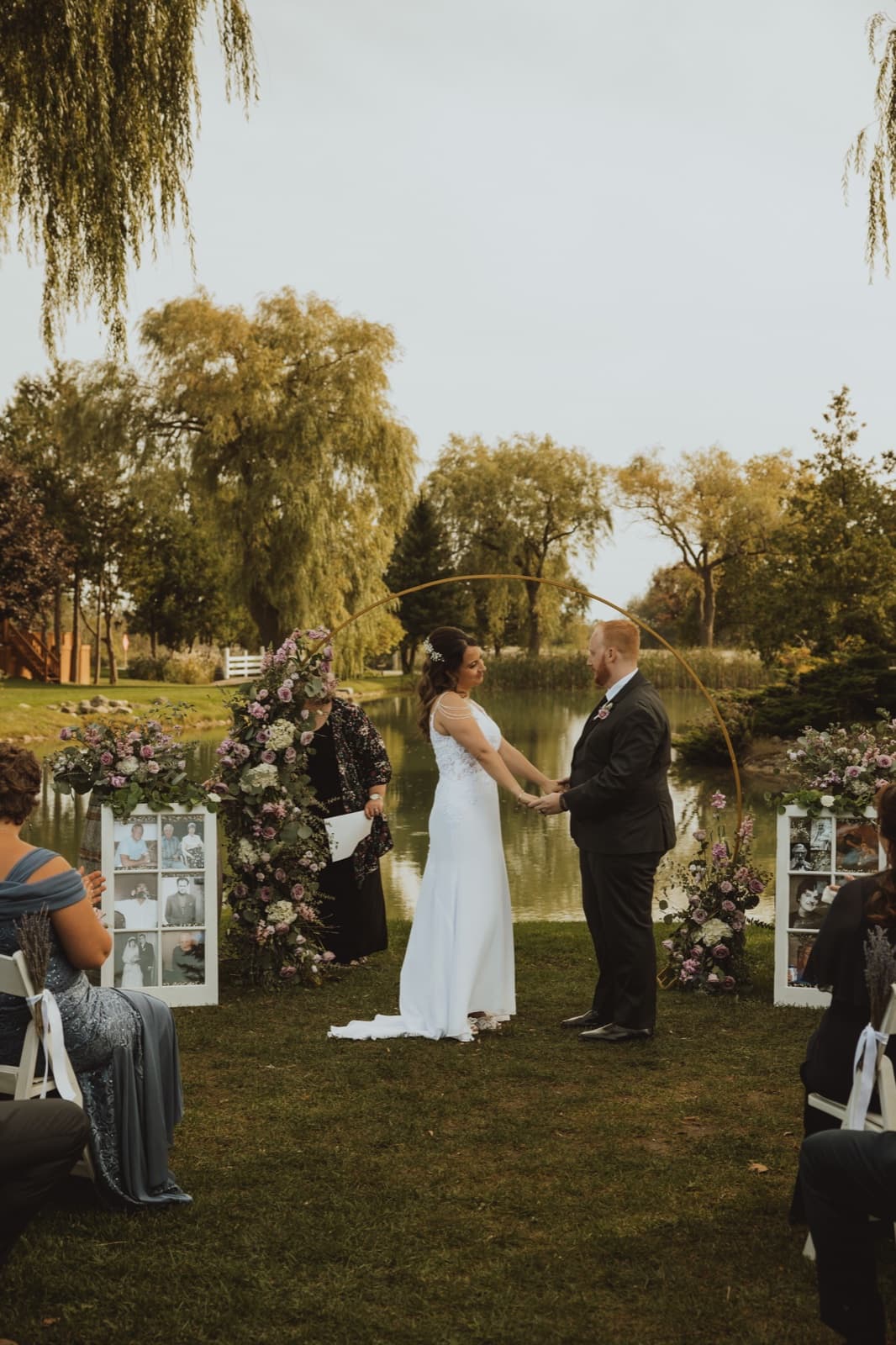 A couple exchanging vows at a gold hoop arch dressed with lilac blooms and greenery beside a pond