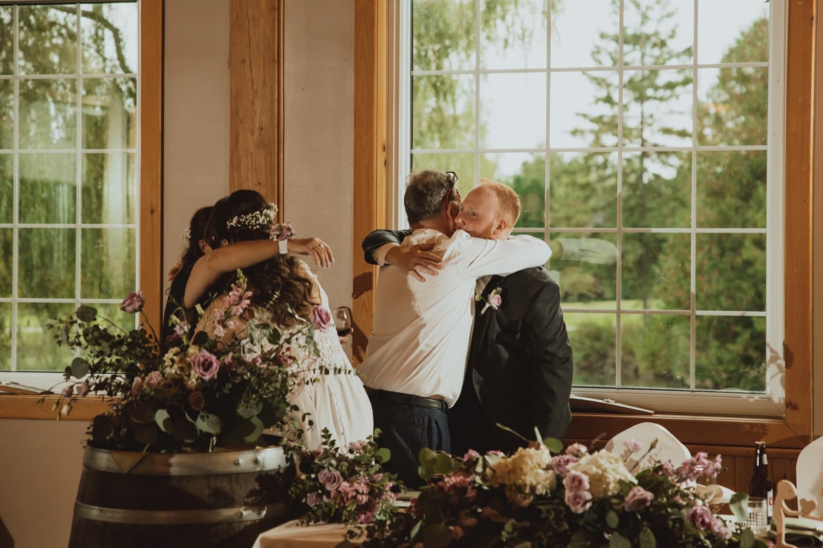 Guests embracing at a reception behind a barrel-top arrangement of mauve roses and eucalyptus
