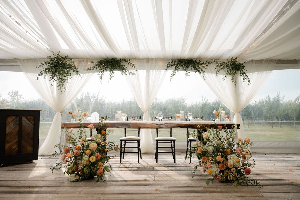 Head table under a white draped tent, flanked by ground arrangements of peach and orange blooms