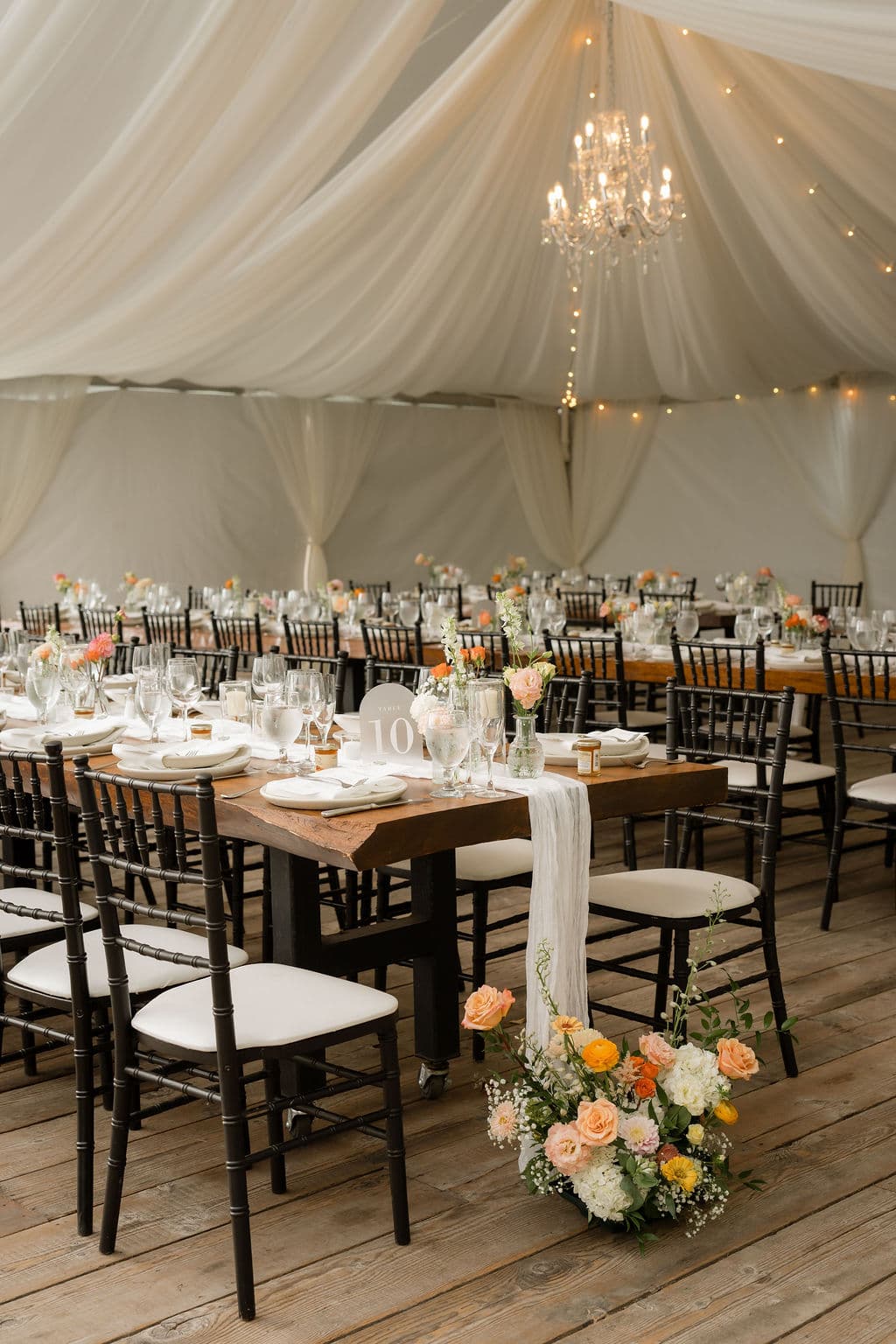 Reception tables under a white draped tent with a chandelier, a peach and white arrangement on the deck