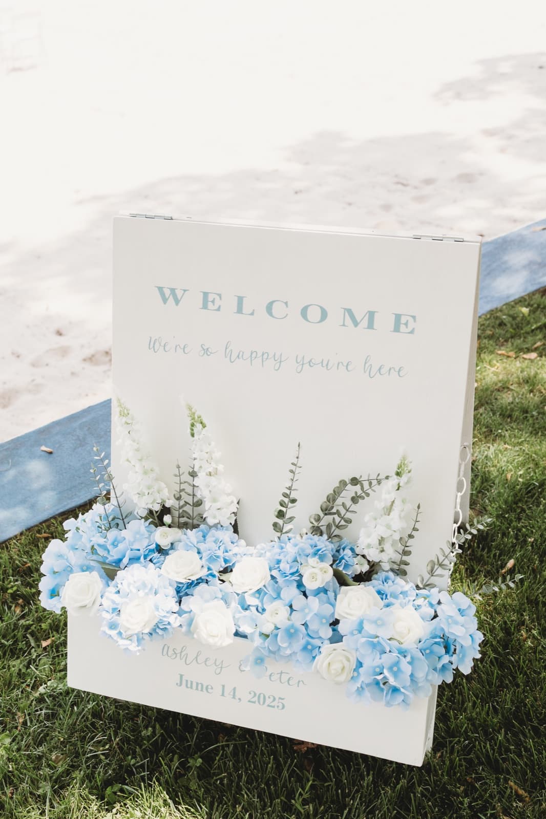 A welcome sign in a planter box filled with blue hydrangea and white roses