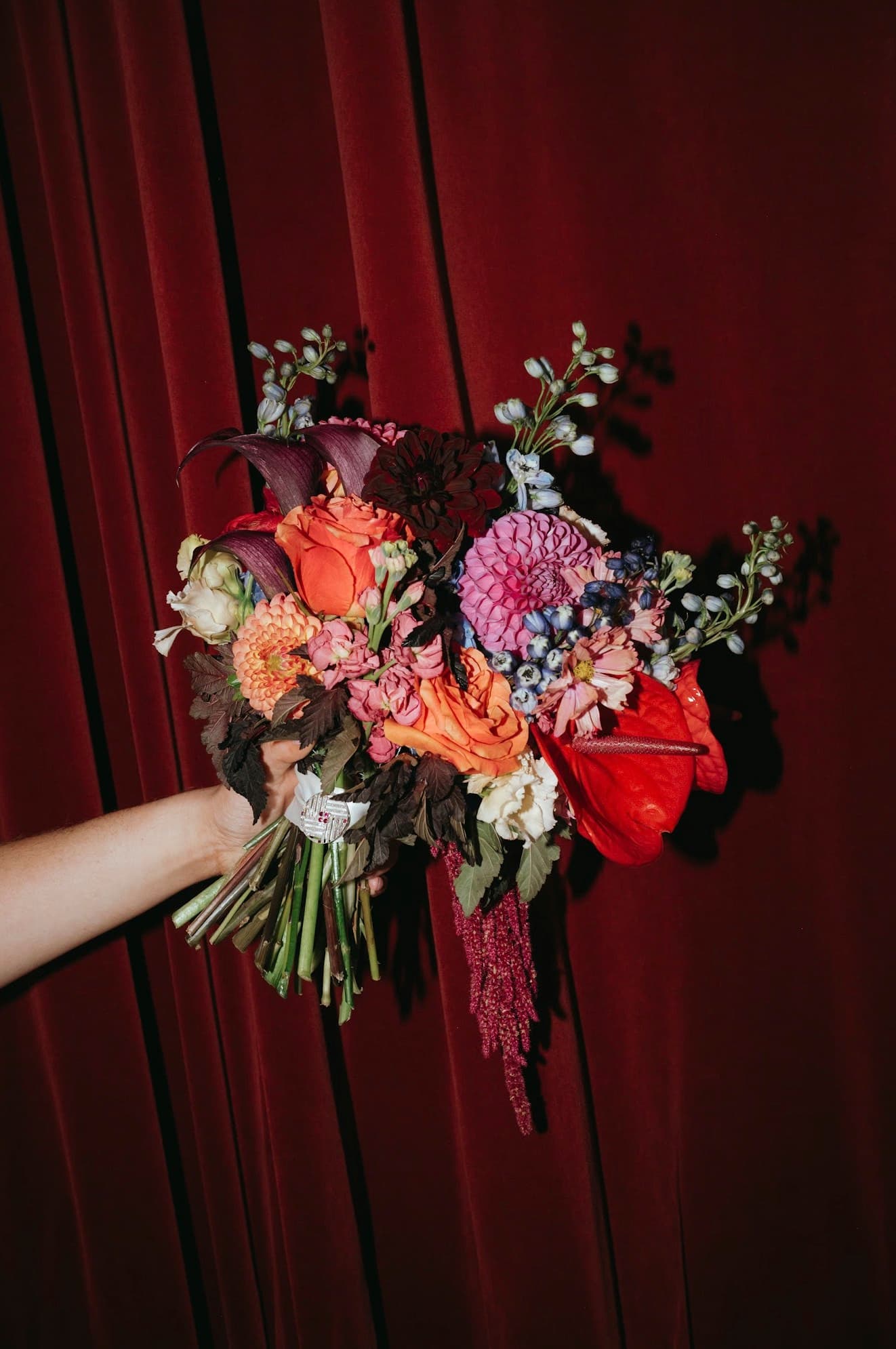 Hand holding a vivid bouquet of orange roses, pink dahlias, anthurium and delphinium against a red velvet curtain